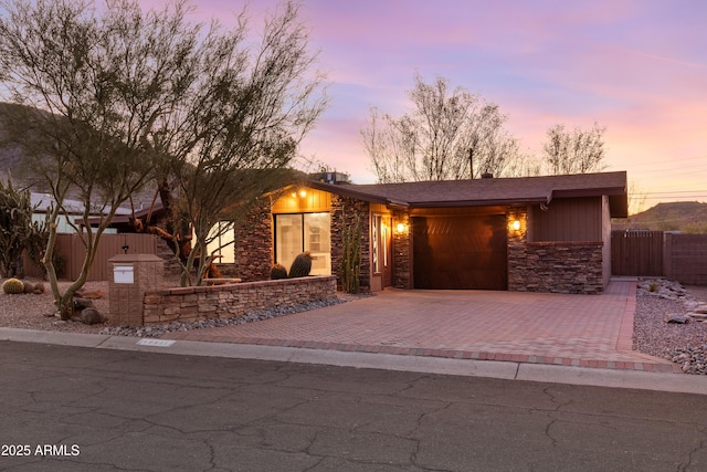 mid-century home with an attached garage, stone siding, fence, and decorative driveway