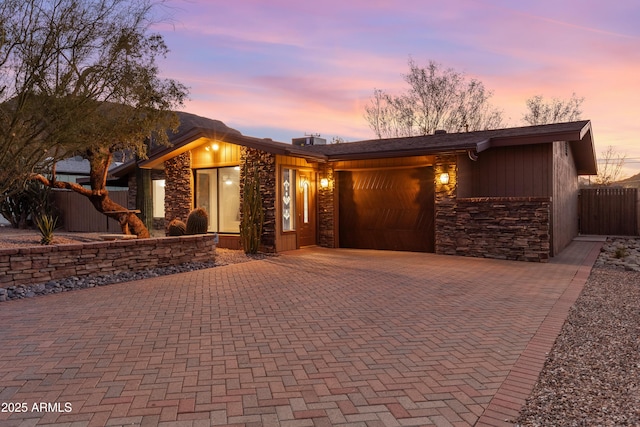 mid-century modern home with stone siding, decorative driveway, an attached garage, and fence