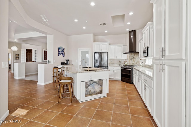 kitchen featuring light tile patterned floors, appliances with stainless steel finishes, wall chimney range hood, a kitchen island with sink, and white cabinets