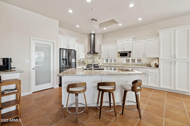 kitchen with white cabinetry, a kitchen island with sink, stainless steel appliances, light stone counters, and wall chimney exhaust hood