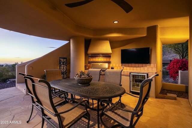 patio terrace at dusk with ceiling fan, beverage cooler, and an outdoor kitchen