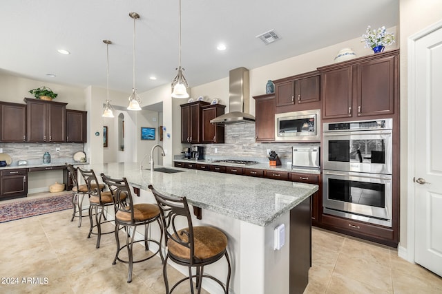 kitchen with light stone countertops, stainless steel appliances, wall chimney range hood, an island with sink, and pendant lighting