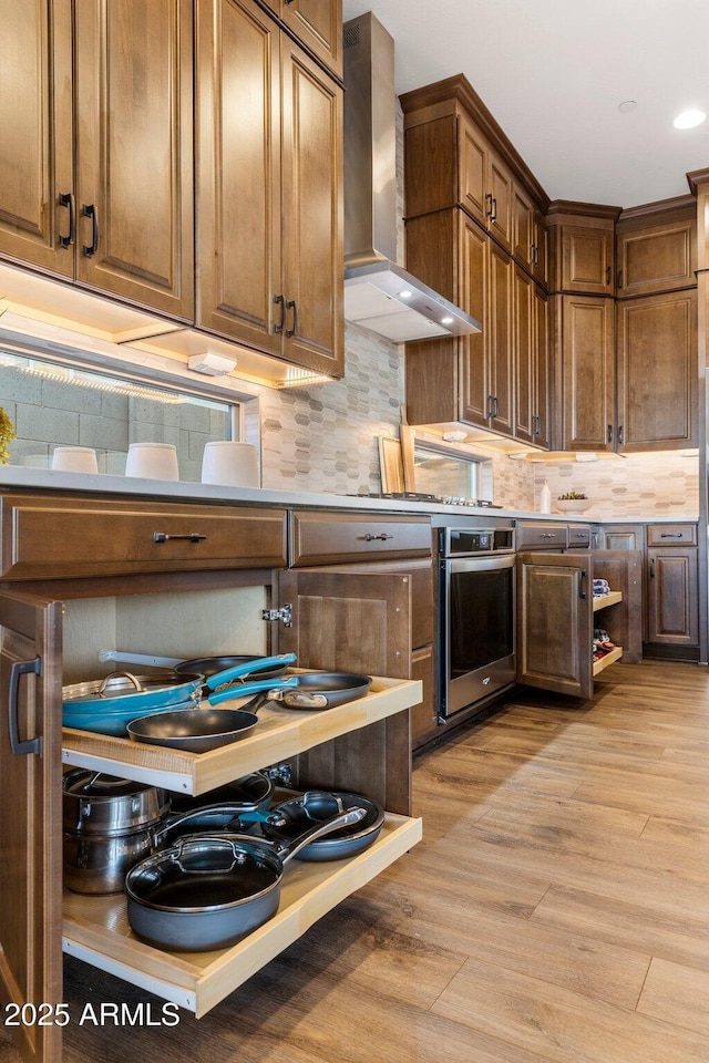 kitchen featuring oven, wall chimney range hood, light wood-type flooring, recessed lighting, and backsplash