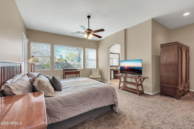 bedroom featuring baseboards, arched walkways, light colored carpet, ensuite bath, and ceiling fan