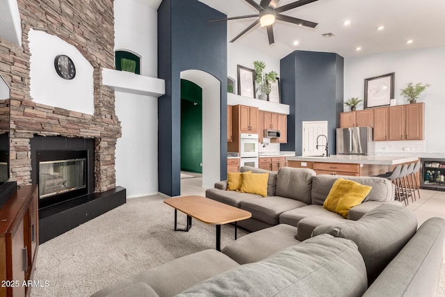 living area featuring a ceiling fan, a towering ceiling, light colored carpet, a stone fireplace, and recessed lighting