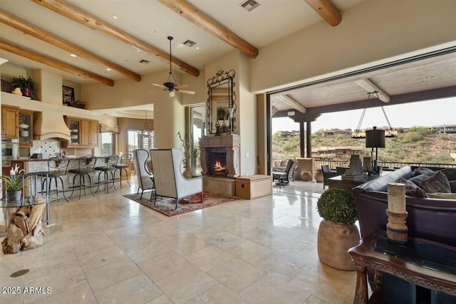 living room featuring a warm lit fireplace, visible vents, beamed ceiling, and ceiling fan with notable chandelier