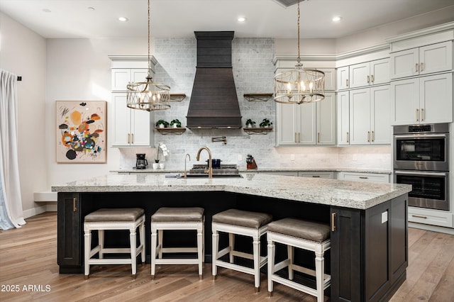 kitchen featuring a chandelier, double oven, a sink, tasteful backsplash, and an island with sink