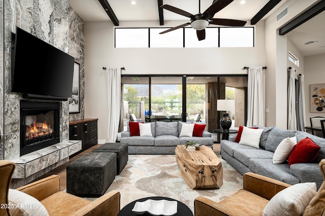 living room with a high end fireplace, a towering ceiling, ceiling fan, beamed ceiling, and plenty of natural light