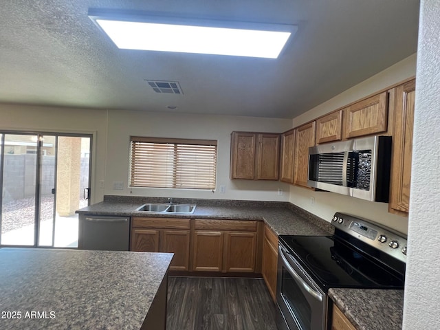 kitchen with appliances with stainless steel finishes, a sink, dark countertops, dark wood-style flooring, and brown cabinets