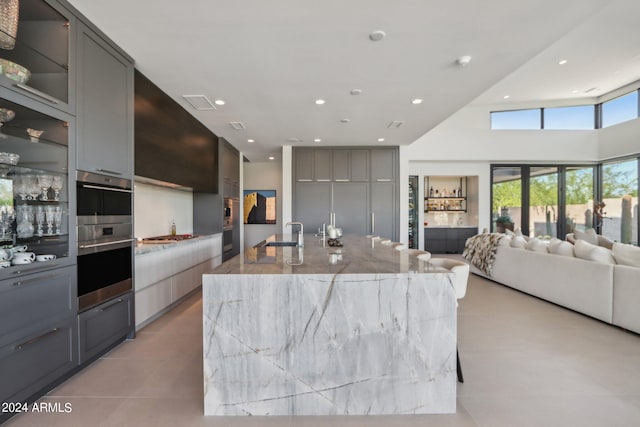 kitchen with a large island with sink, double oven, gray cabinets, a high ceiling, and light stone countertops