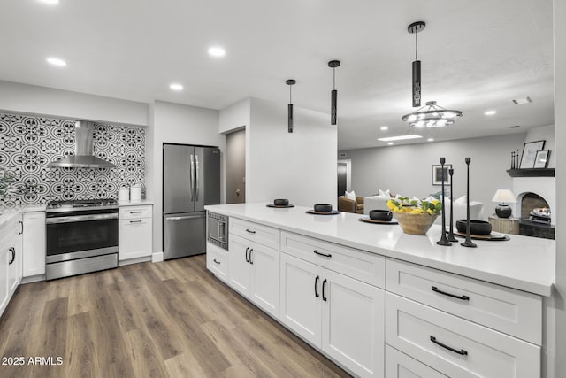 kitchen with wall chimney range hood, light hardwood / wood-style flooring, appliances with stainless steel finishes, white cabinetry, and hanging light fixtures