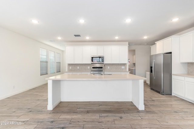 kitchen with an island with sink, backsplash, white cabinets, appliances with stainless steel finishes, and light hardwood / wood-style floors