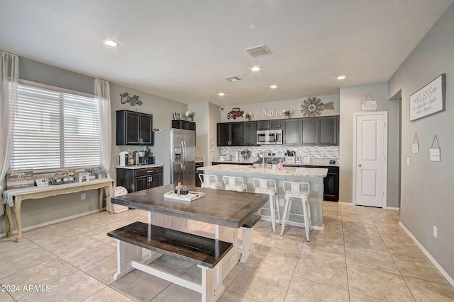 kitchen with visible vents, a kitchen island with sink, backsplash, appliances with stainless steel finishes, and light tile patterned flooring