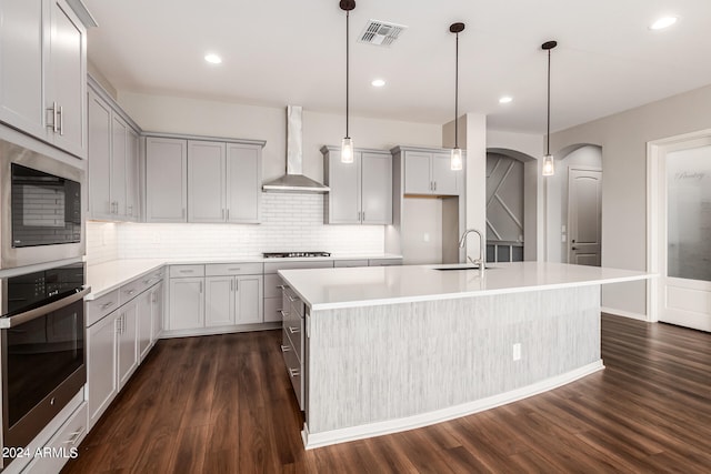 kitchen with dark hardwood / wood-style floors, an island with sink, hanging light fixtures, wall chimney exhaust hood, and appliances with stainless steel finishes