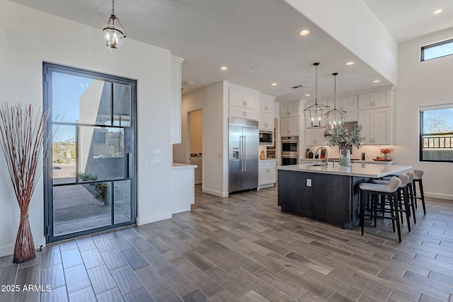 kitchen with decorative light fixtures, white cabinetry, built in appliances, sink, and a center island with sink