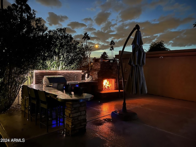 patio terrace at dusk with an outdoor kitchen, an outdoor stone fireplace, an outdoor bar, and grilling area