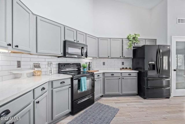 kitchen with backsplash, black appliances, gray cabinets, light wood-type flooring, and a towering ceiling