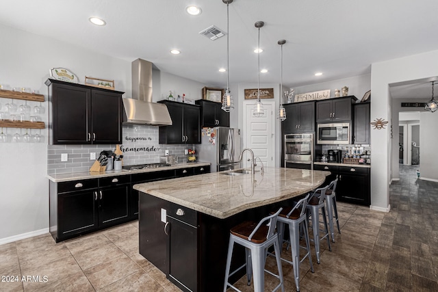 kitchen with decorative backsplash, a kitchen breakfast bar, wall chimney exhaust hood, a kitchen island with sink, and stainless steel appliances