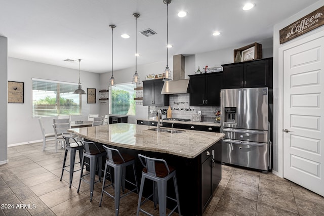 kitchen featuring appliances with stainless steel finishes, light stone counters, a kitchen island with sink, wall chimney range hood, and pendant lighting