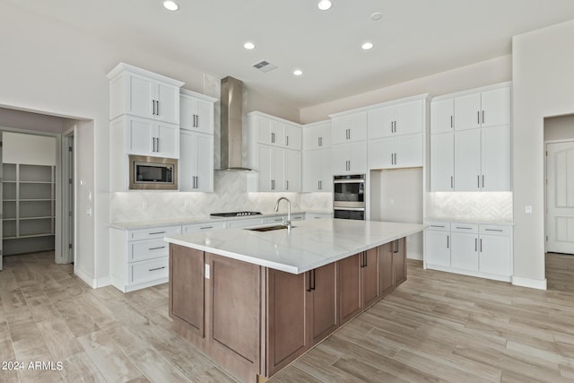 kitchen featuring wall chimney exhaust hood, light stone counters, stainless steel appliances, a kitchen island with sink, and white cabinets