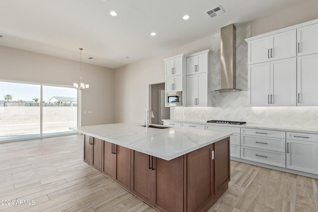 kitchen featuring white cabinets, stainless steel microwave, a kitchen island with sink, and wall chimney range hood