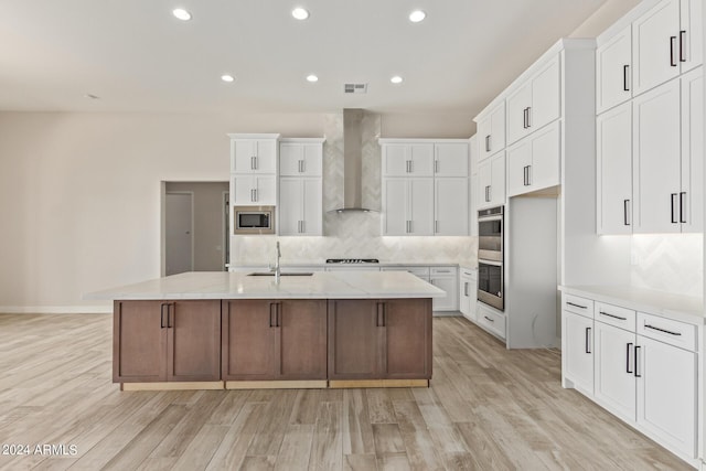 kitchen featuring sink, a center island with sink, appliances with stainless steel finishes, wall chimney range hood, and white cabinets