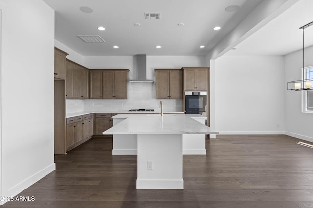 kitchen with light stone counters, visible vents, wall chimney range hood, an island with sink, and dark wood finished floors