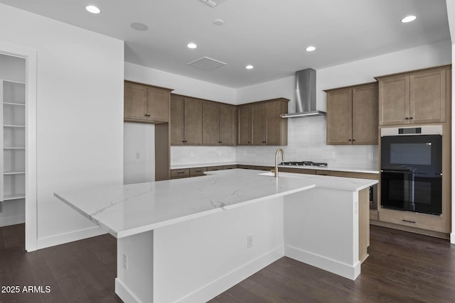 kitchen with dark wood-style floors, a center island with sink, dobule oven black, stainless steel gas stovetop, and wall chimney range hood