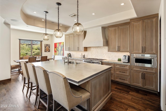 kitchen with premium range hood, a center island with sink, oven, sink, and a tray ceiling