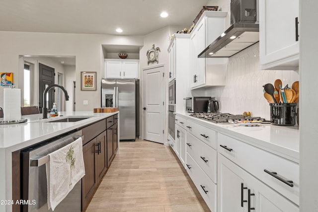 kitchen with appliances with stainless steel finishes, sink, wall chimney range hood, light hardwood / wood-style floors, and white cabinetry