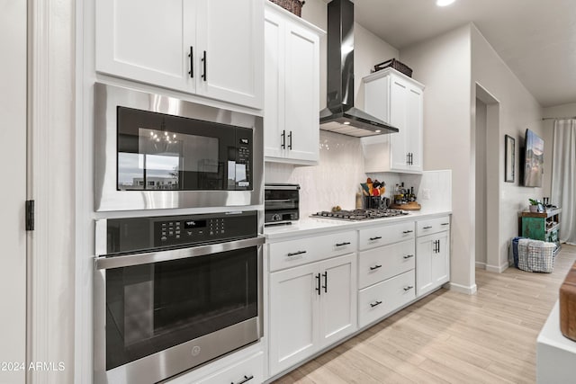 kitchen with wall chimney exhaust hood, decorative backsplash, light wood-type flooring, white cabinetry, and stainless steel appliances