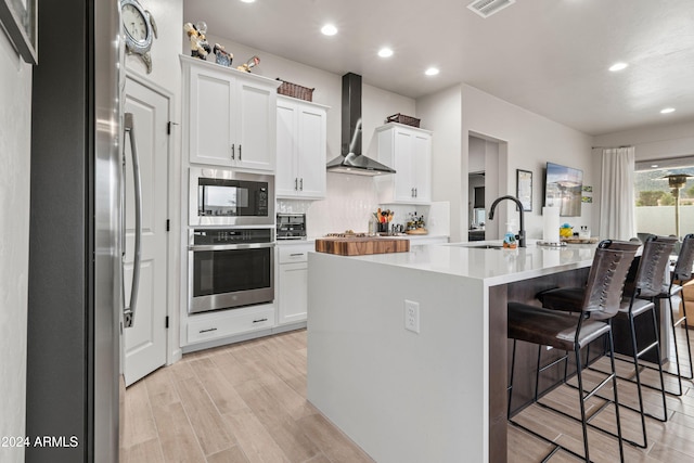 kitchen with stainless steel appliances, white cabinetry, wall chimney exhaust hood, and a center island with sink