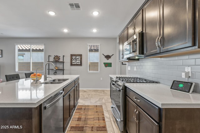 kitchen with dark brown cabinets, appliances with stainless steel finishes, sink, tasteful backsplash, and a kitchen island with sink