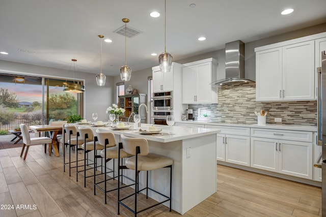 kitchen with white cabinets, a center island with sink, wall chimney exhaust hood, and hanging light fixtures