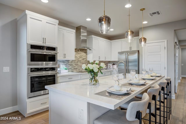 kitchen with light hardwood / wood-style floors, an island with sink, hanging light fixtures, wall chimney exhaust hood, and appliances with stainless steel finishes