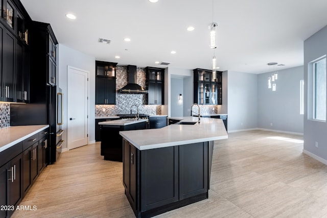 kitchen featuring a kitchen island with sink, wall chimney range hood, sink, hanging light fixtures, and tasteful backsplash