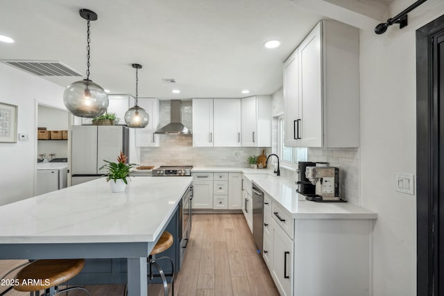 kitchen featuring pendant lighting, white cabinetry, wall chimney range hood, stainless steel appliances, and washing machine and clothes dryer