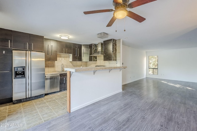 kitchen with wall chimney range hood, a breakfast bar area, backsplash, stainless steel appliances, and dark brown cabinetry