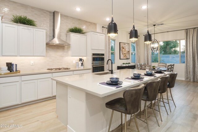 kitchen featuring a center island with sink, light wood-style flooring, stainless steel appliances, light countertops, and wall chimney exhaust hood