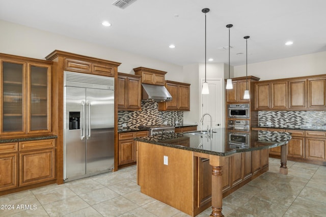 kitchen featuring a kitchen island with sink, exhaust hood, dark stone countertops, appliances with stainless steel finishes, and decorative light fixtures