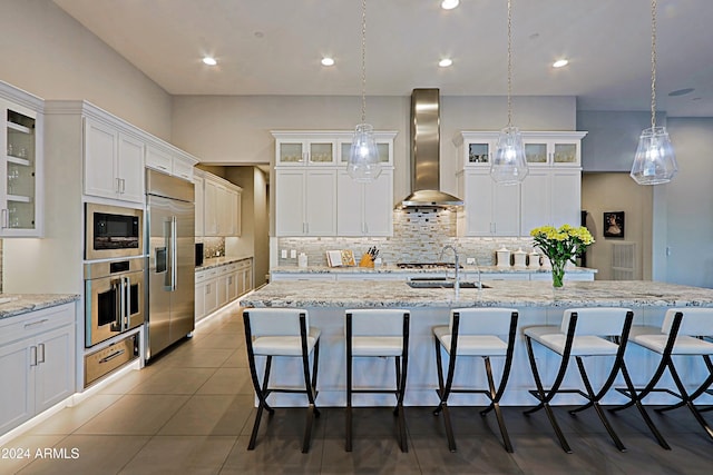 kitchen with wall chimney range hood, built in appliances, a large island with sink, decorative light fixtures, and decorative backsplash