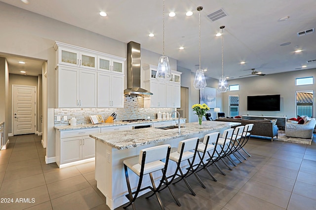 kitchen featuring pendant lighting, wall chimney range hood, sink, an island with sink, and white cabinetry