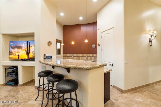 kitchen with kitchen peninsula, light stone countertops, a breakfast bar area, hanging light fixtures, and a towering ceiling