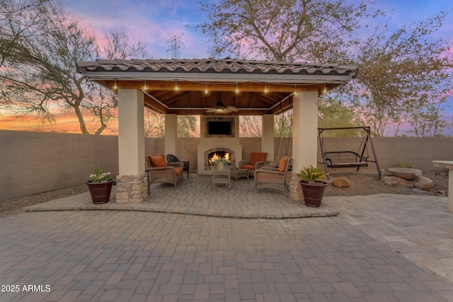 view of patio with a warm lit fireplace, a gazebo, ceiling fan, and a fenced backyard