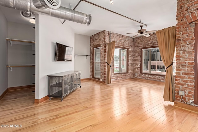 unfurnished living room featuring ceiling fan, brick wall, rail lighting, wood finished floors, and baseboards