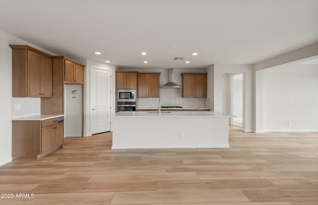 kitchen featuring stainless steel appliances, a kitchen island with sink, light wood-type flooring, and wall chimney exhaust hood