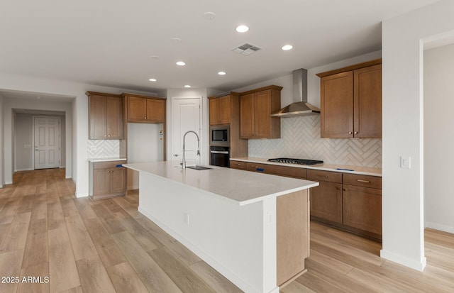 kitchen with sink, a kitchen island with sink, stainless steel appliances, wall chimney exhaust hood, and light wood-type flooring