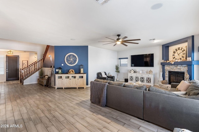 living area with stairway, a ceiling fan, a stone fireplace, light wood-type flooring, and baseboards