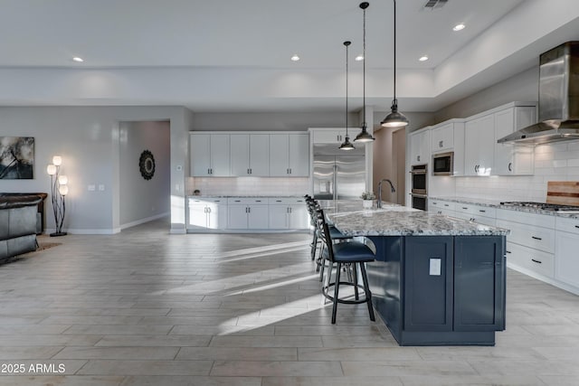 kitchen with pendant lighting, white cabinetry, a large island with sink, built in appliances, and wall chimney exhaust hood