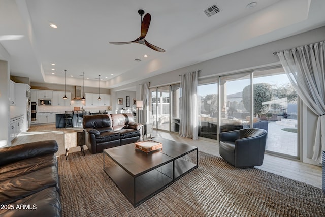 living room with a raised ceiling, ceiling fan, and light wood-type flooring